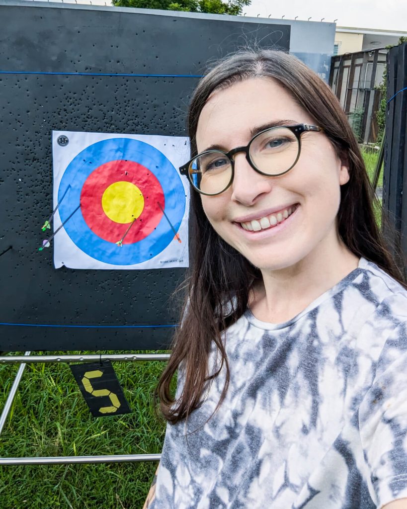 Allison green wearing shorts and a shirt holding a bow and arrow at an archery range in Bangkok