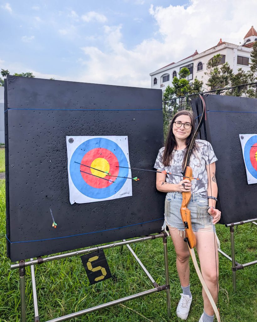 Allison green wearing shorts and a shirt holding a bow and arrow at an archery range in Bangkok