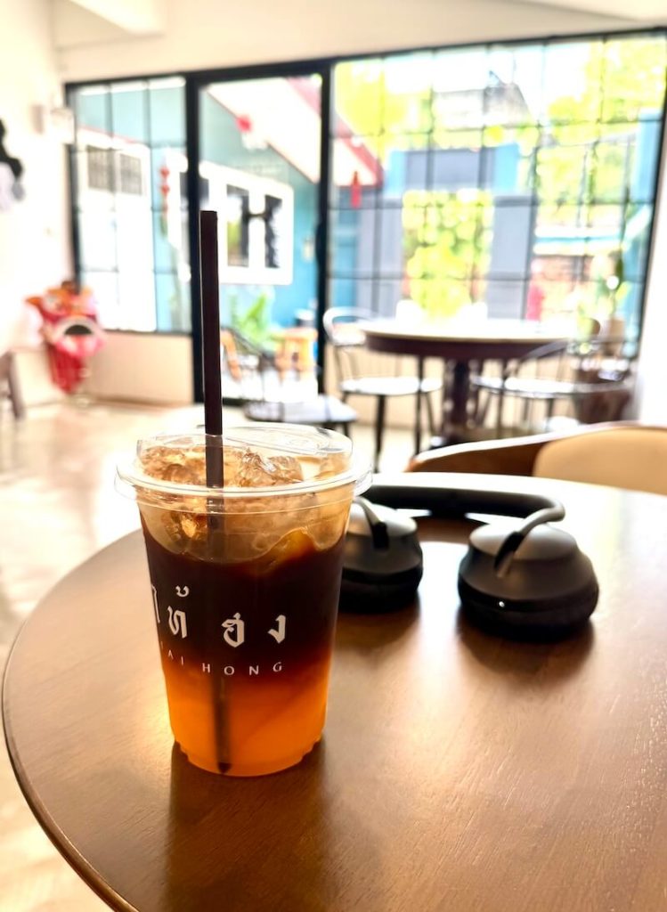 An orange americano in a plastic cup with a straw, in a brightly lit and sparsely populated room, with a pair of headphones on the table.