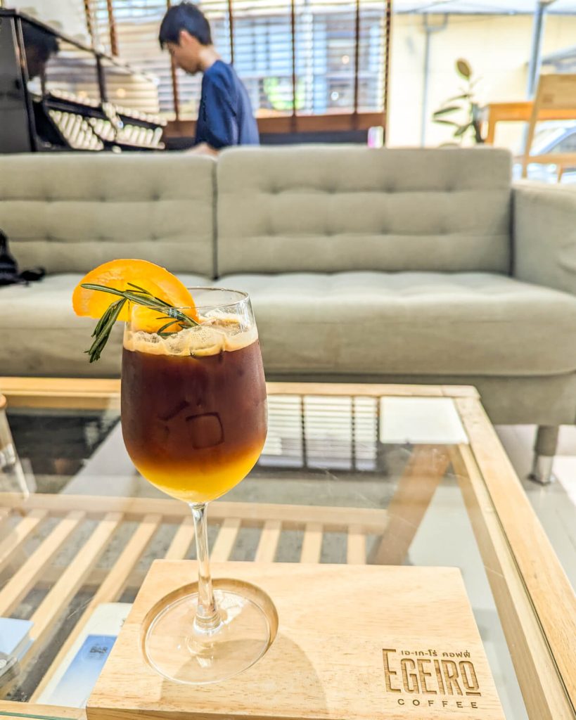 Iced americano drink in a wine glass with rosemary and orange slice with a couch and piano (with a person playing it) in the background